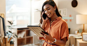an indian woman in an orange tee shirt makes a phone call while looking at a tablet